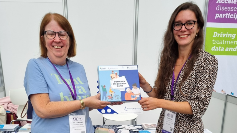 Lewy Body Society CEO Jacqui Cannon in a blue top with Dr Clarissa Giebel in a dress holding the box of the Dementia Inequality Game between them