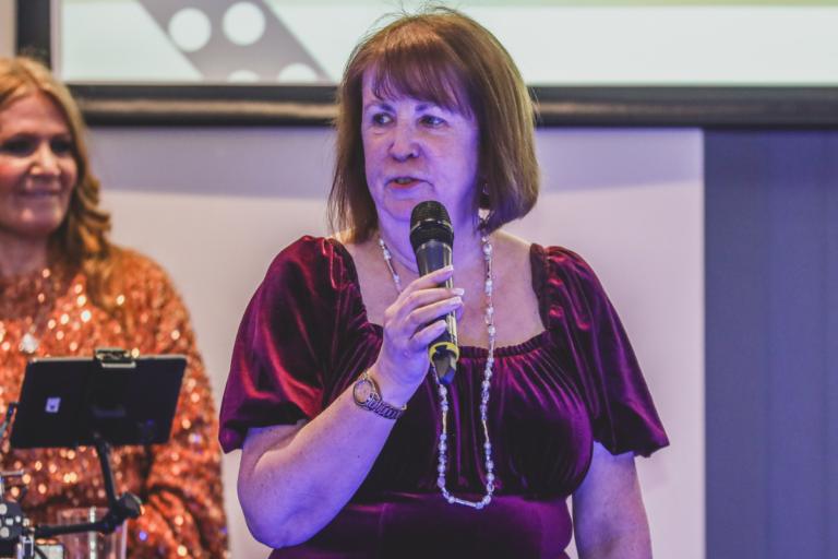 Lewy Body Society CEO Jacqui Cannon - she has brown hair in a bob and it wearing a red dress while holding a microphone