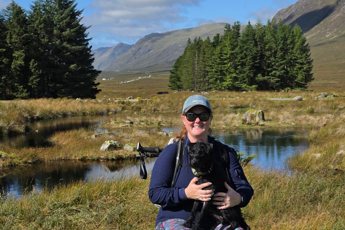 Woman in cap holding black terrier dog in front of some water, trees and mountains on the West Highland Way for Lewy Body Society